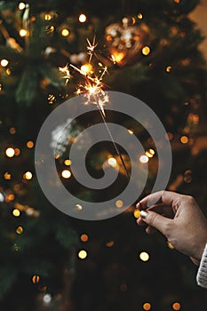 Hand holding firework against christmas tree lights in dark room. Happy New Year! Merry Christmas! Burning sparkler in female hand