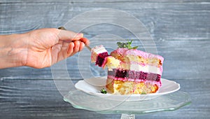 Hand holding a fork with a slice of cake, cleaved from a piece of cake, consisting of pink, white, sand and ruby âEUR