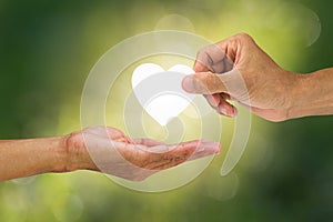 Hand holding and giving white heart to receiving hand on blurred green bokeh background