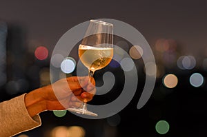 Hand holding a glass of white wine toasting to celebration and party concept on rooftop bar with colorful bokeh of city light and