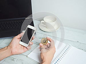 Hand holding gold bitcoin and mobilephone with workspace on white wood table background