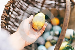 Hand holding golden Easter egg and stylish eggs with green buxus branches in rustic wicker basket on white wooden background.