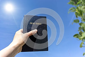 Hand holding Holy Bible against a blue sunny sky