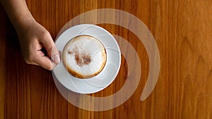 Hand holding hot cappuccino latte in white ceramic cup on wooden background