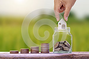 Hand holding house model in a jar for saving money and stacks of coins on a wooden floor, financial concept.