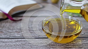 Hand holding jar and pouring out pure virgin olive oil into container with open Bible Book, olives, and green branch