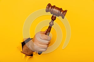 Hand holding a judge's gavel through torn yellow paper wall close-up. Law concept