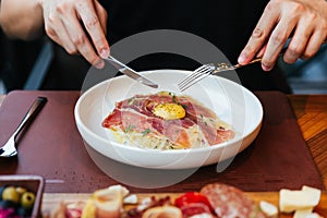 Hand holding knife and fork for eating Fettuccine Carbonara with parma ham and yolk with black pepper. Served in white plate