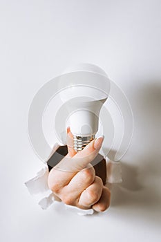 Hand holding a light bulb breaking through the hole in white background.