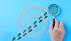 Hand holding magnifying glass focusing on target goal icon on blue background. Business achievement goal and objective target