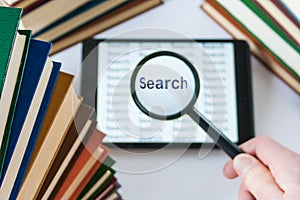 Hand holding a magnifying glass over a tablet surrounded by book