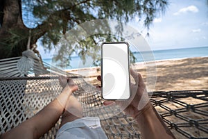 Hand holding mock up mobile with white sceen while laying on hammock swing between trees on the beach