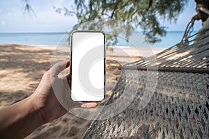 Hand holding mock up mobile with white sceen while laying on hammock swing between trees on the beach