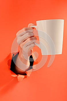 Hand holding a mug of coffee through torn red paper background