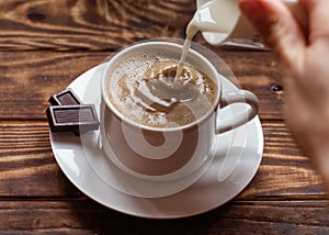 Hand holding mug of milk and pouring it to cap of coffee  on small plate with two small chocolate.   Flat Lay with no people