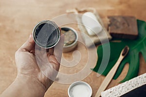 Hand holding natural charcoal toothpaste in glass jar on background of bamboo toothbrush, soap, solid shampoo, metal razor,