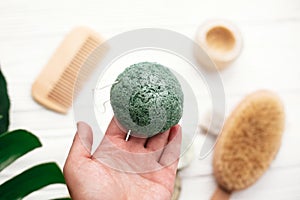 Hand holding natural konjaku sponge on background of bamboo brush, deodorant in glass on white wood with green monstera leaves. Z photo