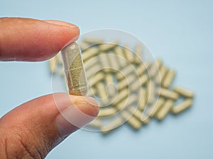 Hand holding of one Andrographis paniculata capsule with a pile of capsules  on blue background.