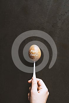 Hand holding one Happy Easter written egg in a spoon on black metal background