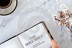 Hand holding open holy bible book, cup of coffee, and spring branch with white flowers