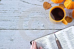 Hand holding open holy bible book of psalms with coffee cup, autumn leaves, and pumpkin on wooden table, top view