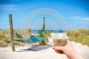 Hand holding paper cup of hot coffee. Relax with empty hammock and ocean background on tropical beach, vacation concept