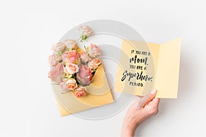 hand holding paper with mothers day phrase beside pink flowers in envelope