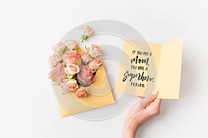 hand holding paper with mothers day phrase beside pink flowers in envelope isolated on white