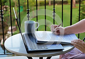 hand holding pen over paperwork with laptop on balcony