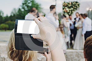 Hand holding phone with empty screen and taking photo of gorgeous bride and stylish groom with guests at wedding reception in