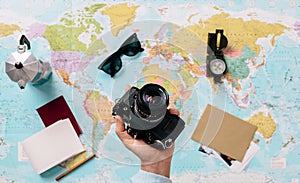 Hand holding a photo camera with a world map in the background next to a compass, sunglasses, notepad, compass and an Italian