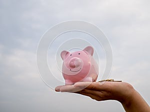 Hand holding pink piggy bank on the blue sky with cloud background, Saving money for future plan and retirement fund concept