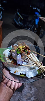 hand holding a plate of food, namely chicken satay and rice cake