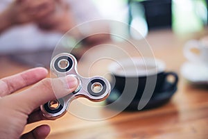 A hand holding and playing metal silver color fidget spinner with coffee cups and people using mobile phone