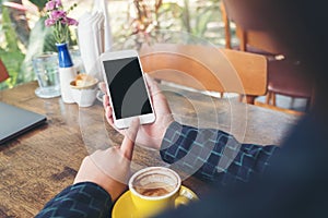 A hand holding and pointing at white mobile phone with blank black desktop screen with yellow coffee cup on wooden table in cafe