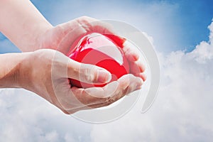 Hand holding red hart on blue sky and white clouds, with bright light