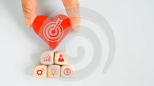 Hand holding red heart with dartboard above business icon on wood cube for target or goal concept