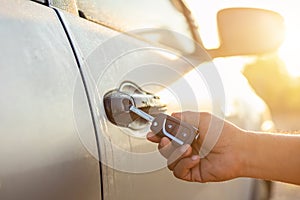 Hand holding remote car keys and press the button open or lock the silver car at outdoor parking lot with sunlight effect in