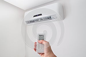 Hand holding remote control for air conditioner on white wall.