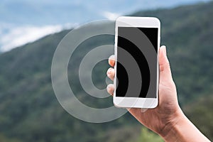 A hand holding and showing white smart phone with blank black desktop screen in outdoor with blur green mountains background