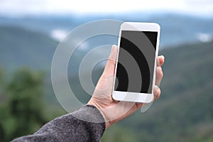 A hand holding and showing white smart phone with blank desktop screen in outdoor with blur green mountains background