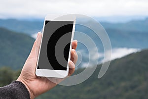 A hand holding and showing white smart phone with blank desktop screen in outdoor with blur green mountains background