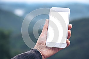 A hand holding and showing white smart phone with blank desktop screen in outdoor with blur green mountains background