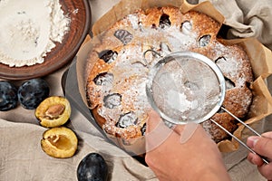 A hand holding sieve sieving sugar powder on plum pie. Plum cake decoration with sugar icing