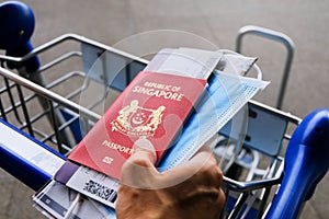 Hand holding Singapore passport, mask and boarding pass, holding on to airport trolley