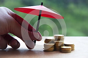 Hand holding small red umbrella over pile of gold coins on wooden table. Financial safety and investment concept.