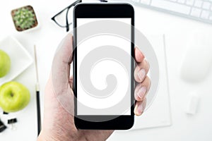 Hand is holding a smartphone with blank mockup screen above the white desk table