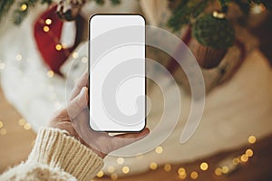 Hand holding smartphone with empty screen against stylish festive christmas gifts and golden lights. Christmas phone mock up.