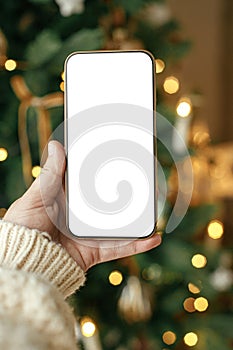 Hand holding smartphone with empty screen against stylish festive christmas tree with golden lights.Christmas phone mock up. Space