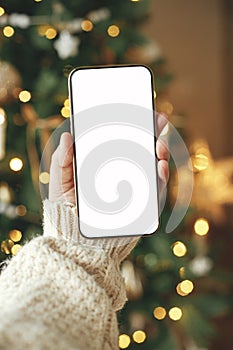 Hand holding smartphone with empty screen against stylish festive christmas tree with golden lights.Christmas phone mock up. Space